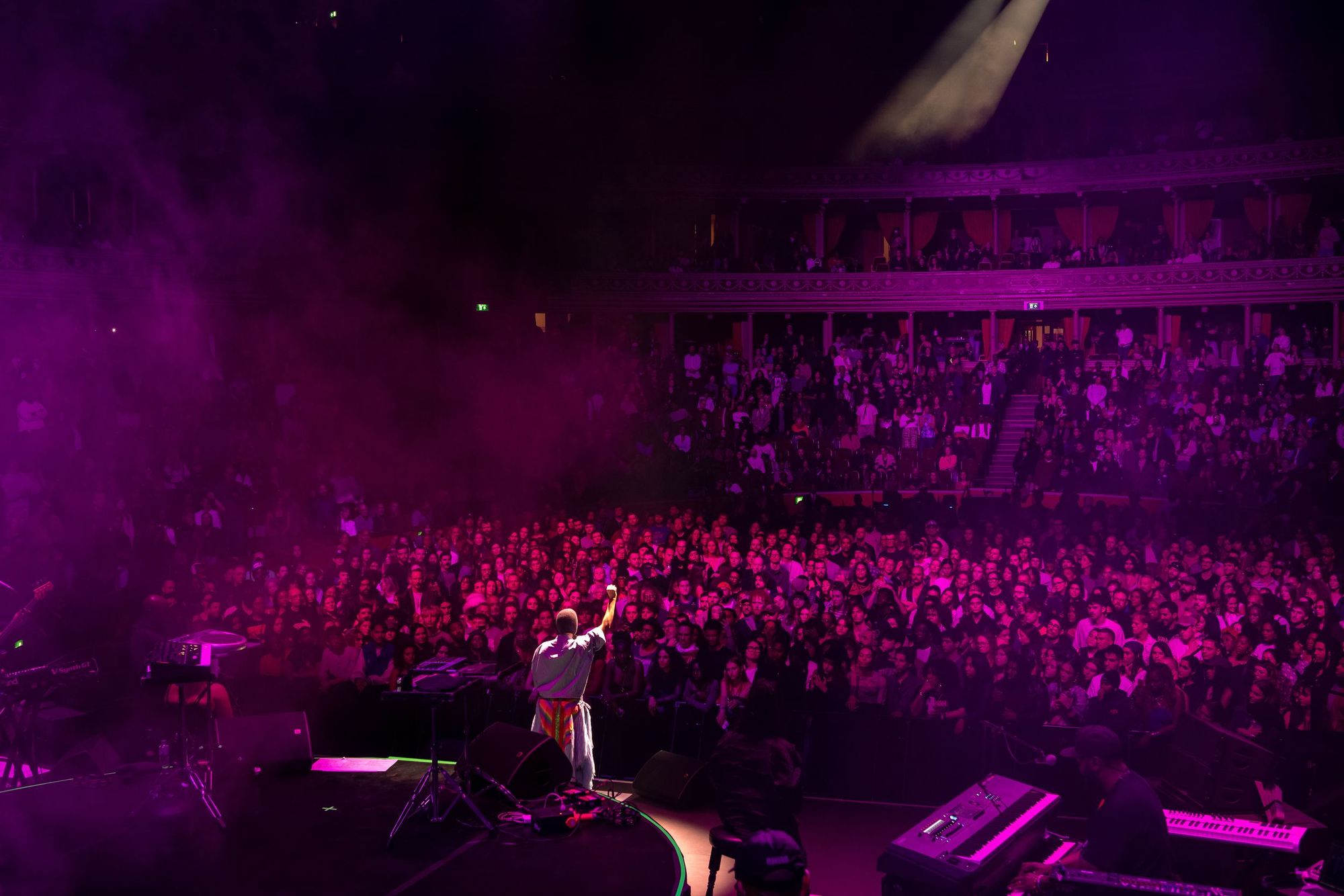 A full concert hall seen from the back, with the stage lit in purple