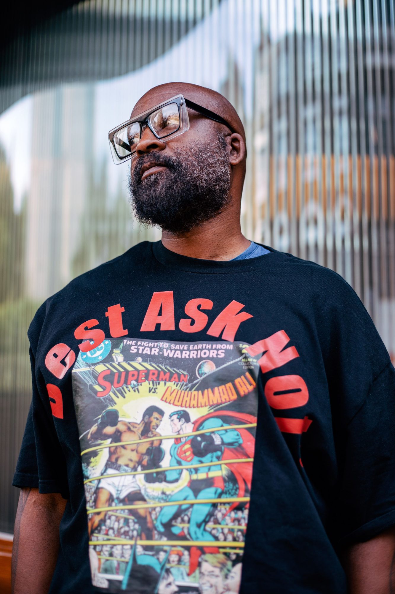 Man in glasses and a comic print t-shirt looking up, glass building behind