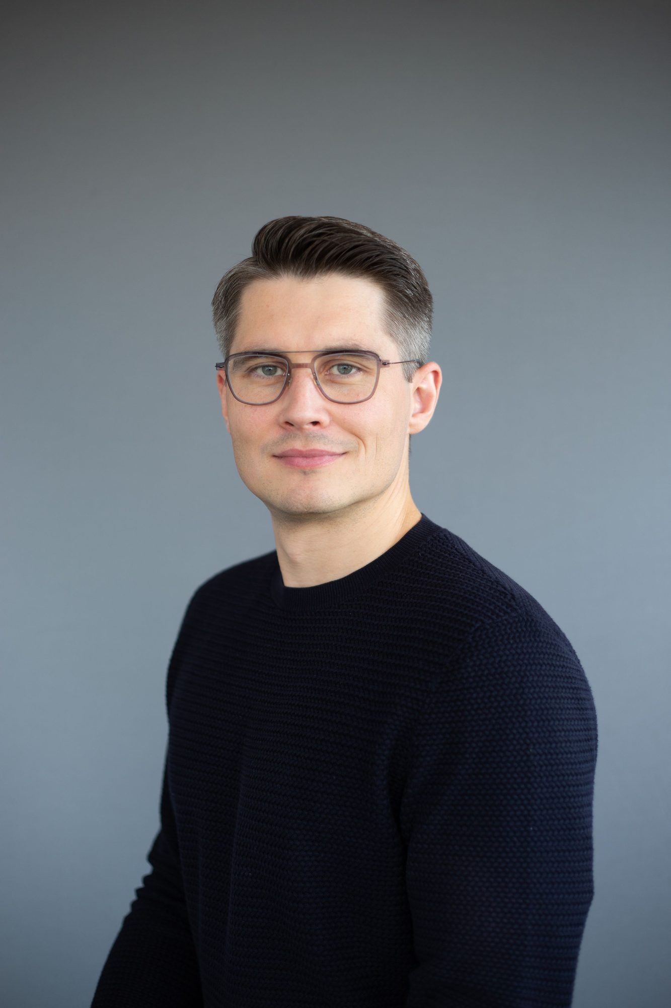 Man in glasses and a navy jumper against a grey backdrop