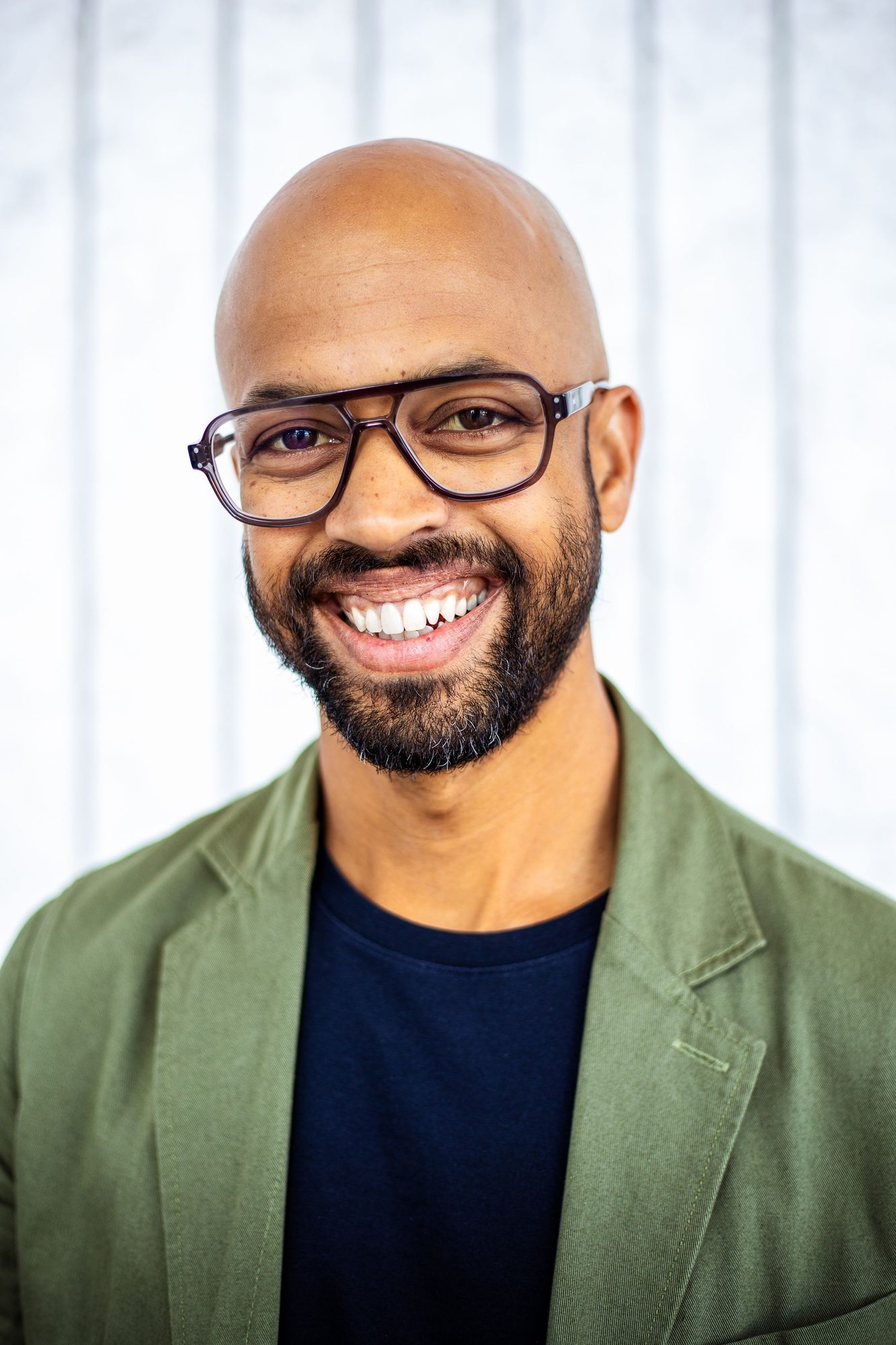 Smiling man in glasses and a green jacket against a white panelled wall