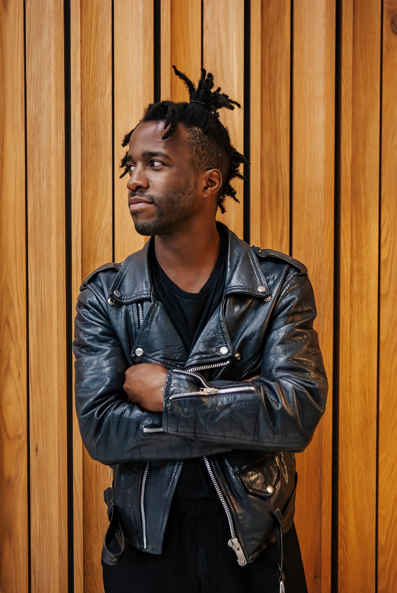 Man with locs in a black leather jacket, arms folded against a timber wall
