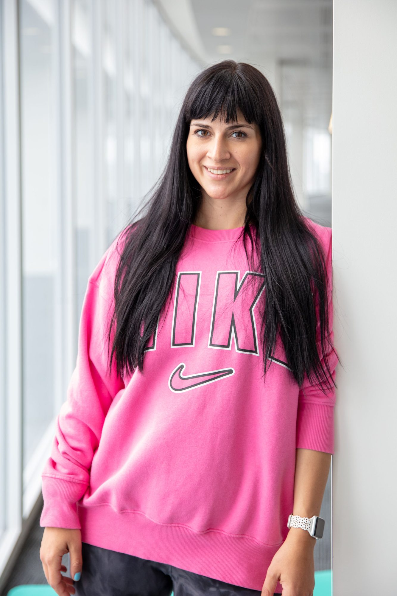 Woman with long dark hair in a pink sweatshirt, standing by a bright office window