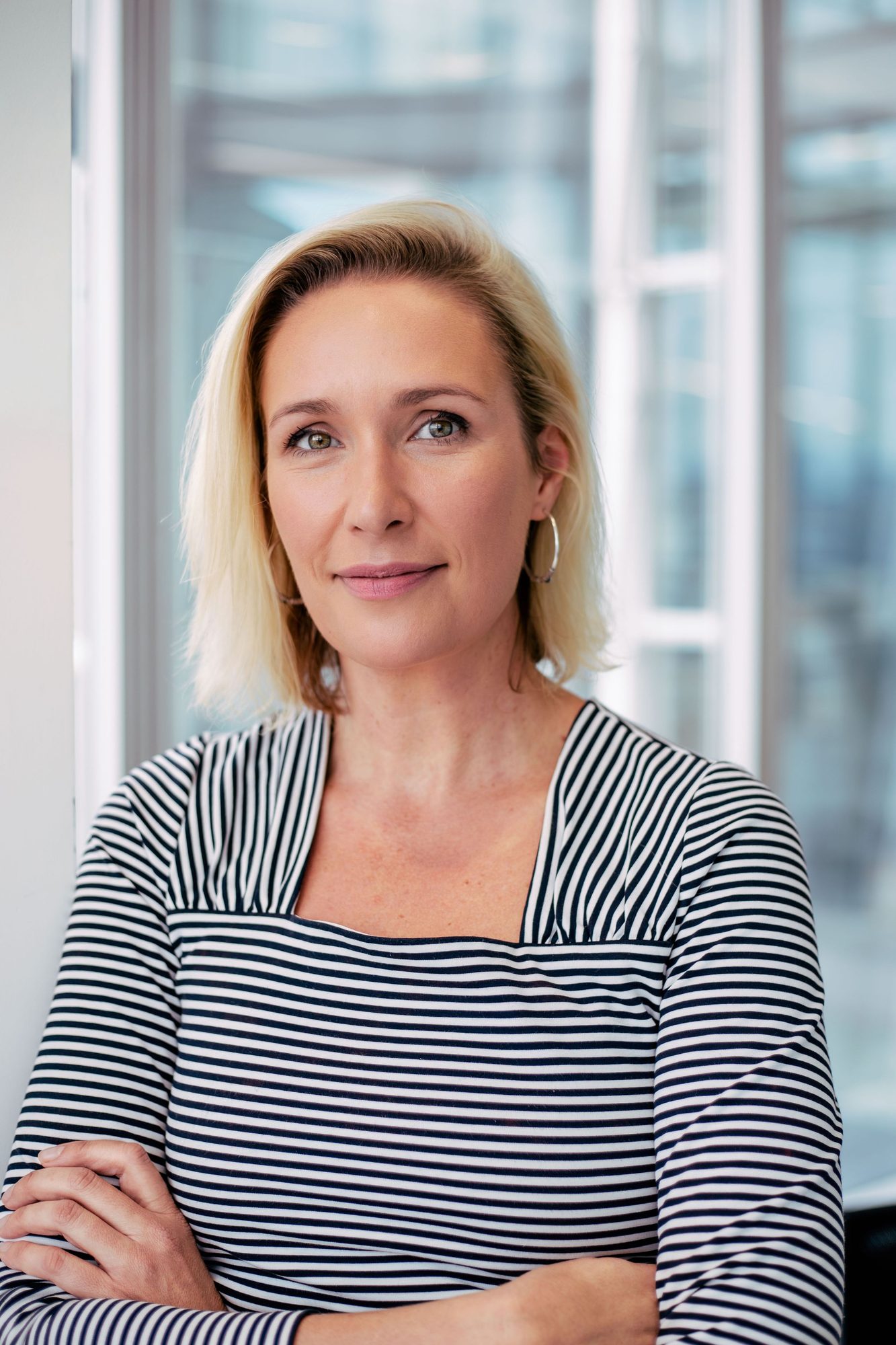Blonde woman in a striped top with arms folded by a window